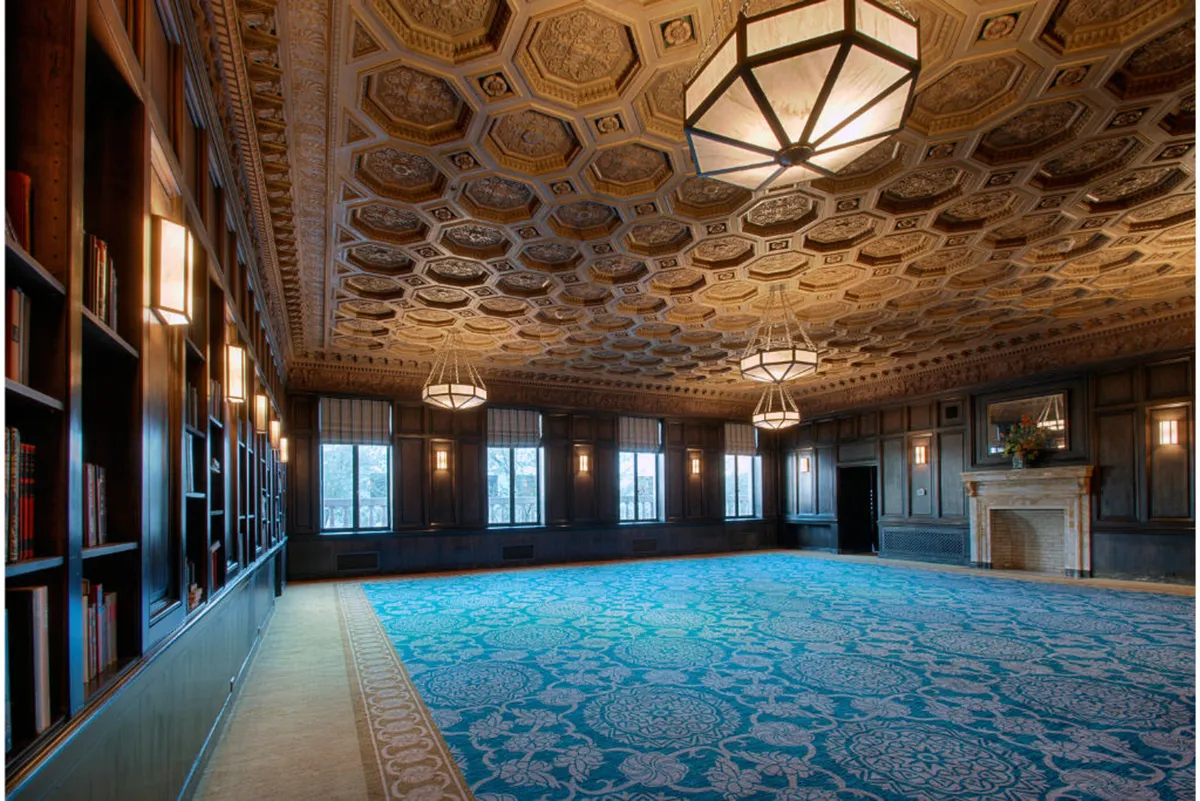 A sophisticated interior with an ornate ceiling, bookshelves filled with literature, a fireplace, decorative light fixtures, and expansive windows letting in natural light. The room feels welcoming, intellectual, and conducive to both creative work and social gatherings.