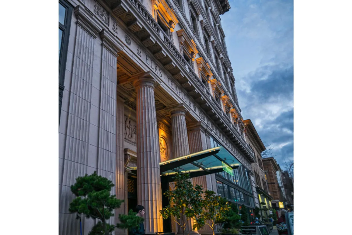 An exterior view showcasing ornate columns, glowing light fixtures, greenery, and a glass entrance. The scene captures a lively outdoor atmosphere suitable for dining and socializing, with hints of vibrant city life and upscale ambiance.