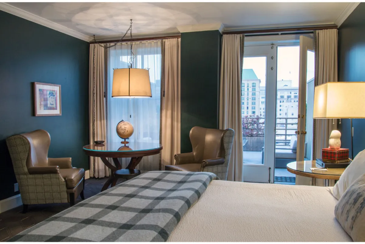 A refined bedroom corner with a comfortable bed draped in a plaid throw, a globe resting on a side table, a modern hanging light fixture, and a pair of inviting chairs. The room is softly lit, suggesting a space for relaxation, reading, or enjoying a morning beverage.
