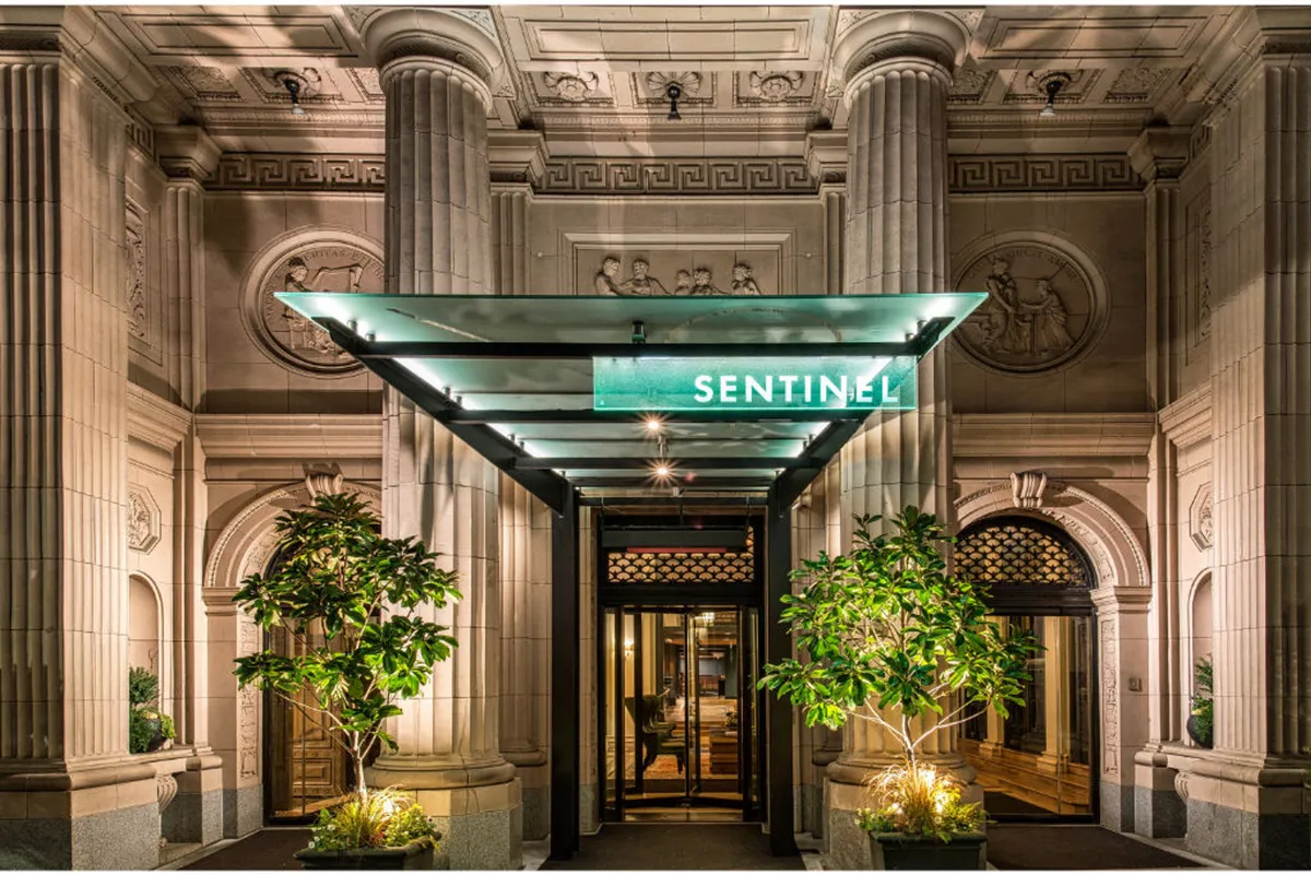 The elegant entrance of the Sentinel property, with ornate architectural details, lush planters, and a prominent awning bearing the 'SENTINEL' name. The scene is inviting, sophisticated, and hints at the exclusive experience awaiting inside.