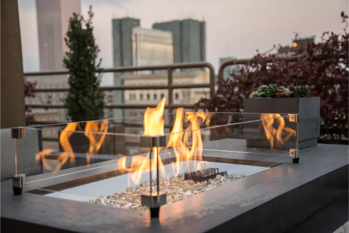 A private outdoor terrace featuring a modern fire pit, cozy seating area, glass enclosure, and a captivating city skyline visible in the background. The evening ambiance is warm and intimate, with the fire pit glowing as dusk settles over the city.