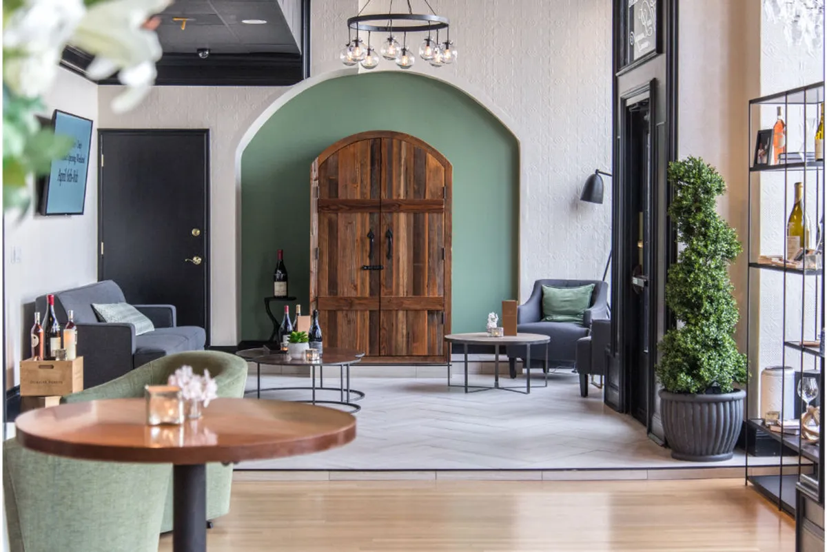 A stylish and inviting space with a green accent wall, wooden door, circular chandelier, and a comfortable seating arrangement. Shelves with bottles hint at wine service, while the setup encourages casual gatherings, wine tastings, and relaxed conversations.