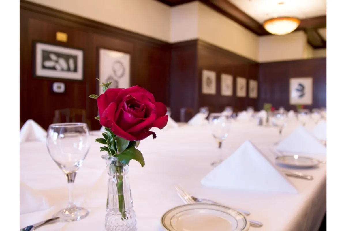 An elegantly set dining table adorned with a single red rose, framed art on the walls, and soft, romantic lighting. The ambiance is intimate and sophisticated, suggesting a private and secluded dining experience ideal for couples.