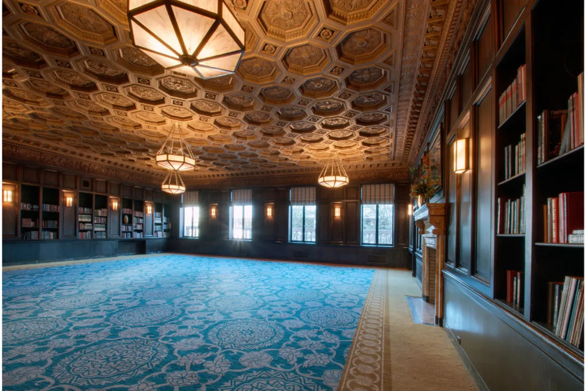 A regal, inviting room with ornate ceiling details, large windows letting in natural light, bookshelves lining the walls, and a blue patterned carpet. The space is designed for reading, social gatherings, and meetings, supporting both focused work and networking.