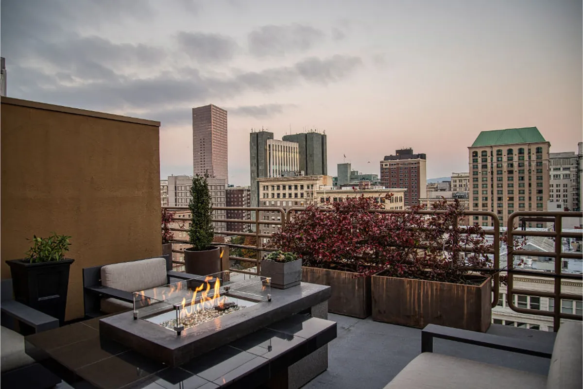 The image showcases a sophisticated rooftop terrace at Hotel Sentinel, featuring a glowing fire pit surrounded by modern seating, lush planters brimming with greenery, and sweeping views of Portland’s sparkling city skyline at dusk. The serene ambiance invites guests to unwind or gather for memorable evenings high above the city.