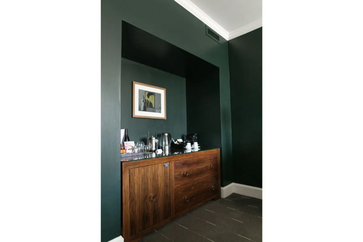 A cozy coffee station set atop a wooden cabinet, complete with a gourmet coffee setup, cups, and premium tea. Framed photographic art adorns the wall, spotlighting the room's artistic touches and local flair.