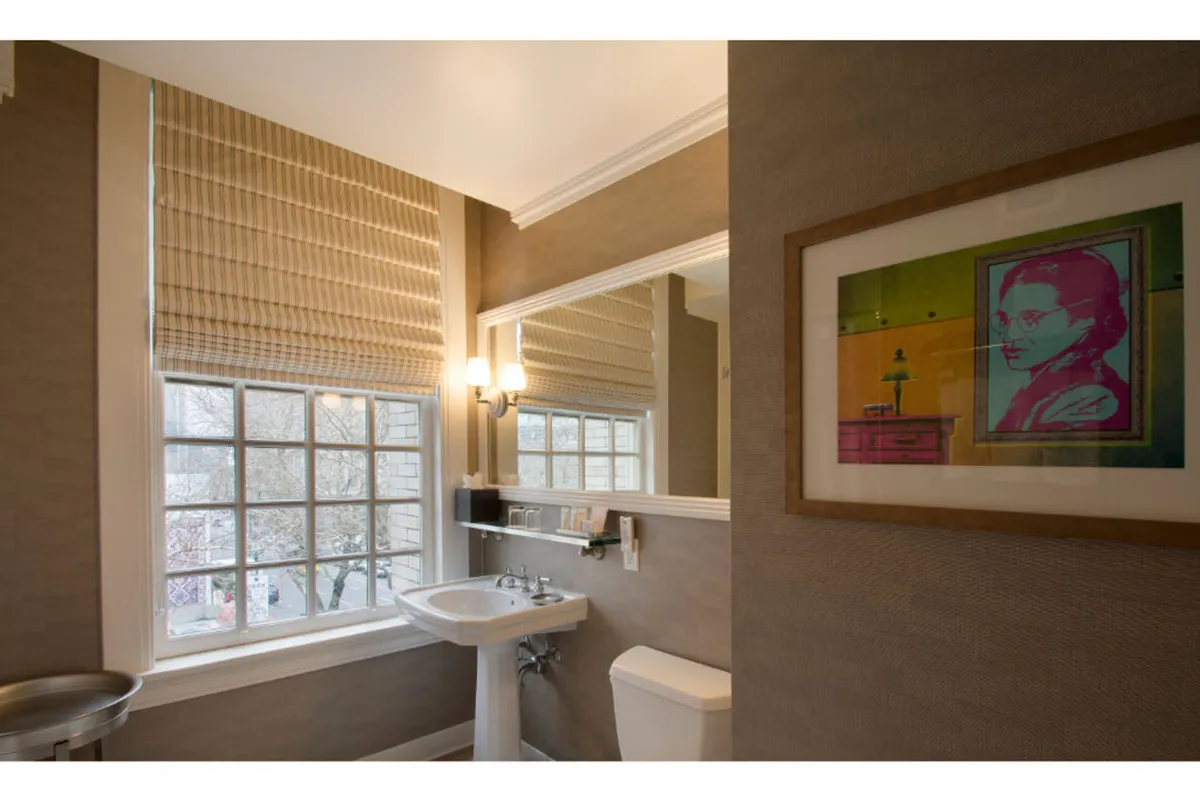 A contemporary bathroom scene featuring a window with blinds, a modern sink, a mirror, sconce lighting, and framed artwork. The space feels fresh, artistic, and welcoming, allowing guests to enjoy moments of self-care and style.