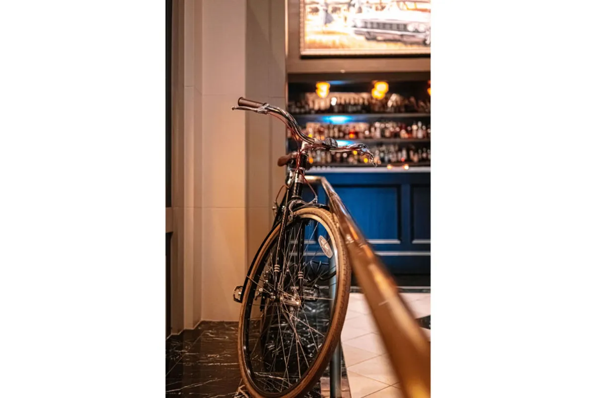 A casual, inviting bar setting with a prominent bicycle near the bar counter, warmly lit by contemporary lighting fixtures. The presence of the bicycle subtly connects the space to active, wellness-focused urban exploration, while the bar invites informal networking or post-meeting relaxation.
