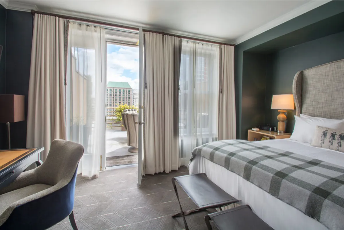 A serene bedroom scene featuring a plush bed topped with a plaid blanket, open balcony doors letting in natural light, a decorative lamp on a side table, and a writing desk set against the wall. The space exudes comfort and sophistication, ideal for both restful nights and productive work sessions.