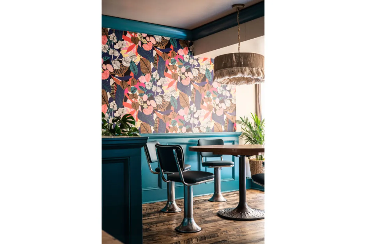 A cheerful dining space with floral patterned wallpaper, a wooden table, metal chairs, and a hanging light fixture. The scene feels open and inviting, with a relaxed ambiance ideal for socializing or unwinding after a city stroll.