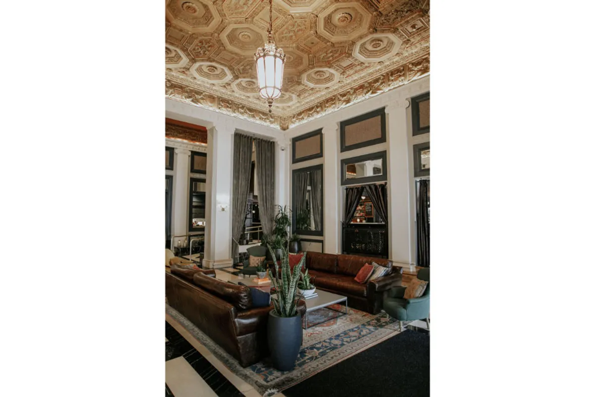 A sophisticated lounge featuring a high ornate ceiling, plush and luxurious seating, a large potted plant, and a decorative rug. The atmosphere is both cozy and elegant, with guests seen relaxing, socializing, and reading, all framed by soft evening lighting.